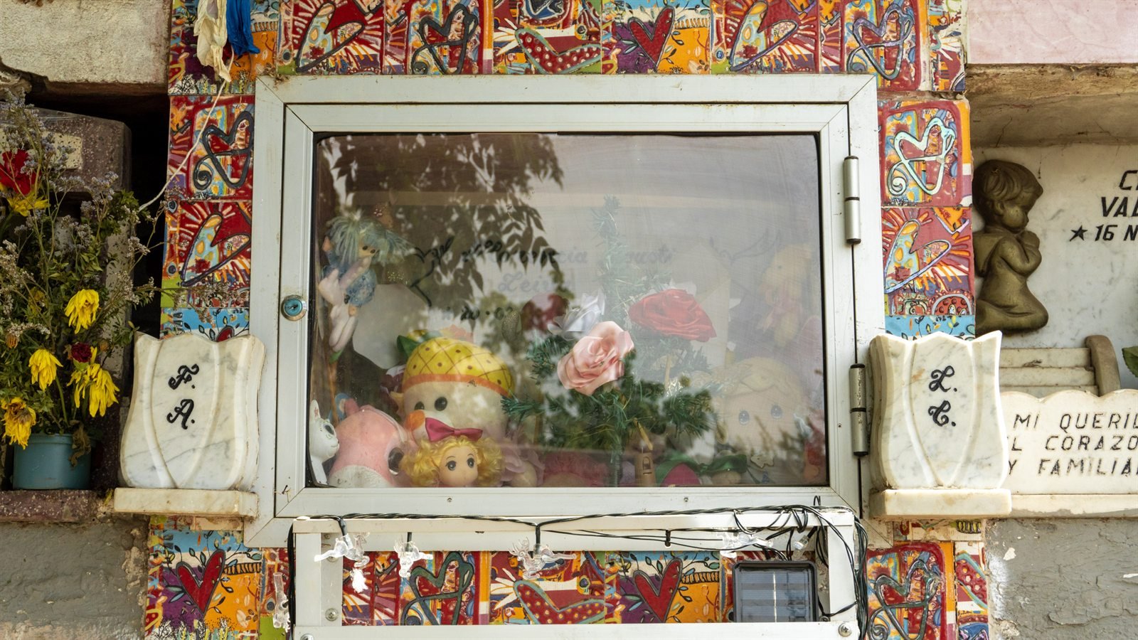 Nicho funerario decorado con azulejos de corazones, flores y muñecos dentro de una vitrina.
