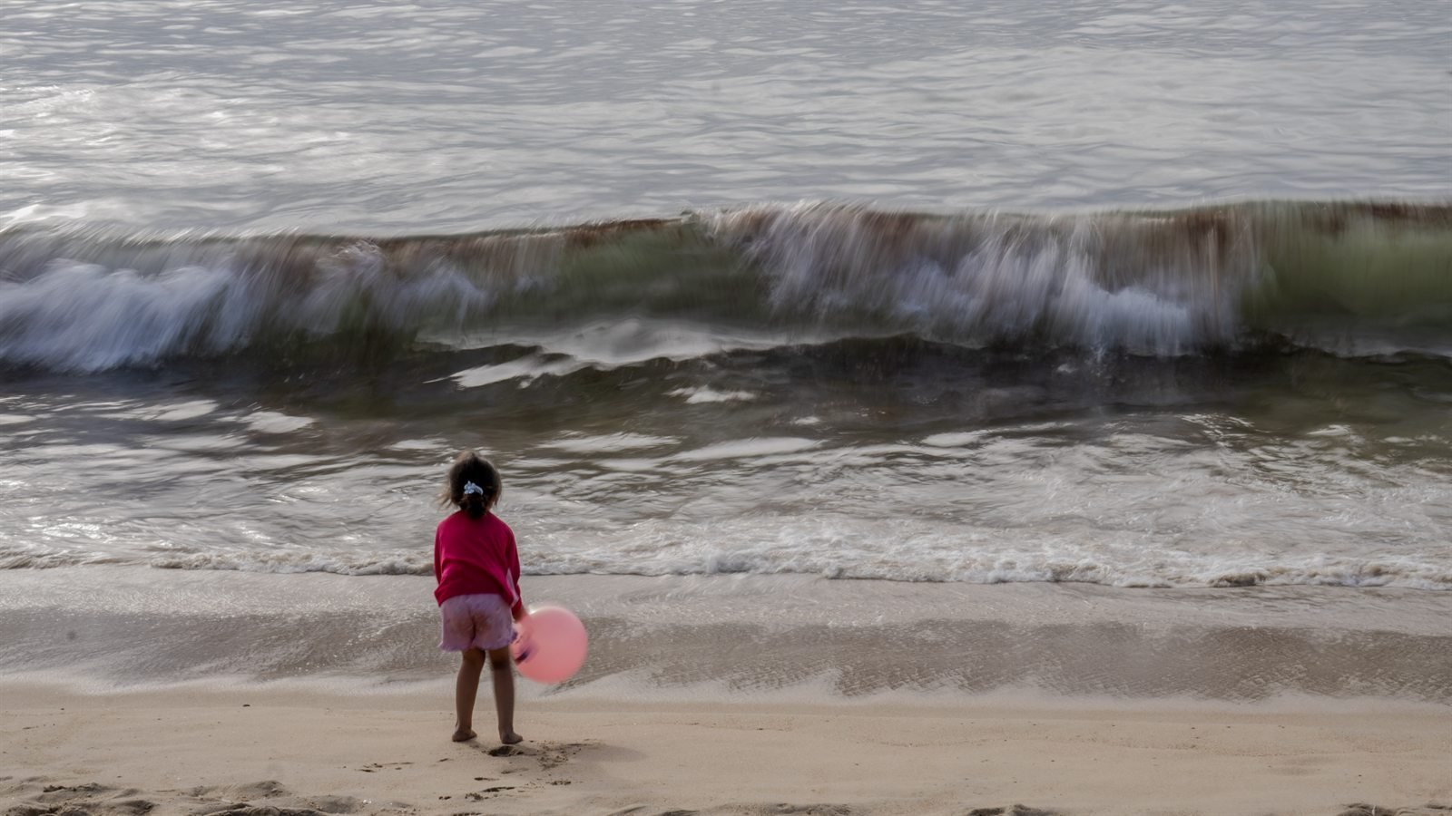 Niño de espaldas con un globo rosa observa las olas romper en la playa.